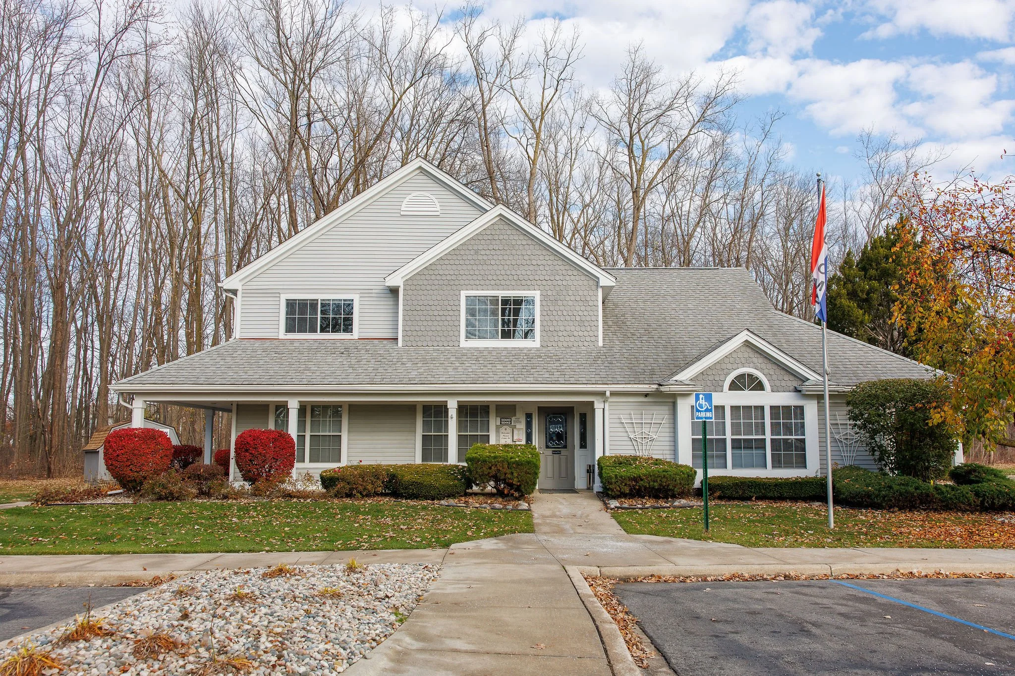 White multifamily residential building with a porch, surrounded by bushes and trees, a flag, and a parking sign outside.