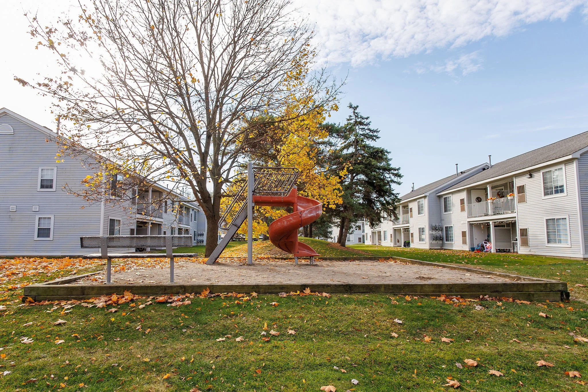 A playground with a spiral slide, a bench, and a sandpit area surrounded by residential apartment buildings. Fall leaves are scattered on the grass and sand, with trees displaying autumn colors under a partly cloudy sky.