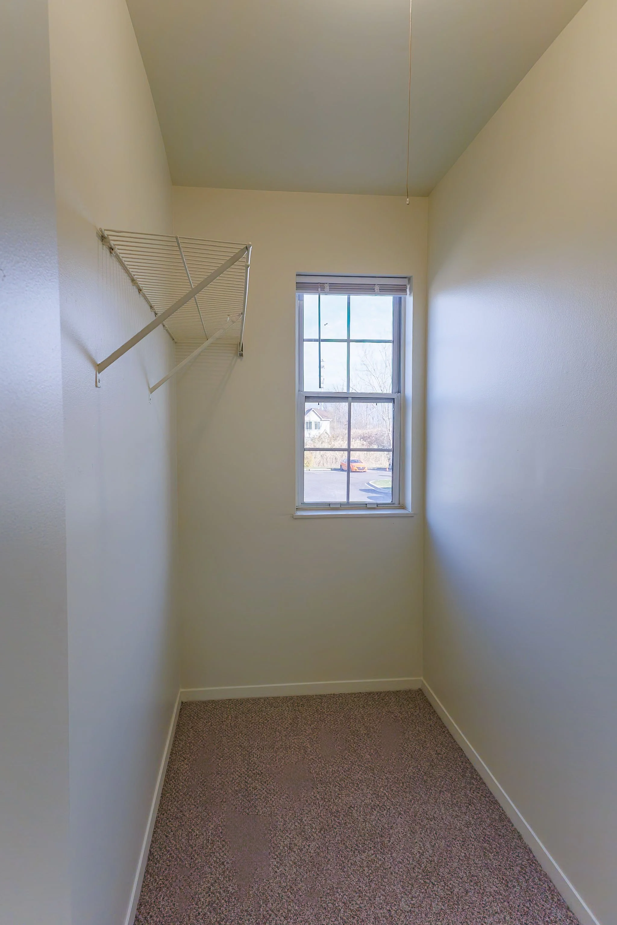 Small empty walk-in closet with a window, white walls, beige carpet, and a wire shelf on the left wall.