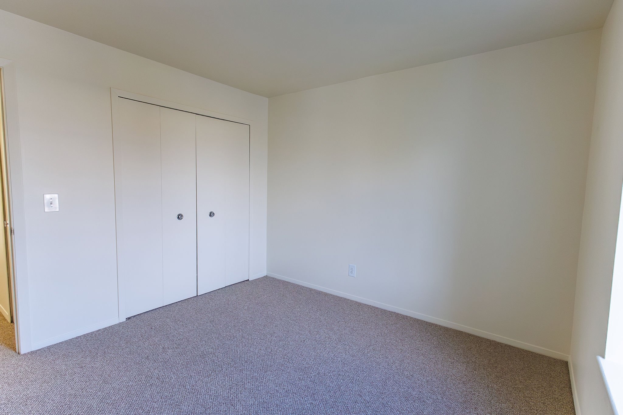 Empty room with beige carpet, white walls, a closet with two doors, a window on the right side, and a white electrical outlet on the wall.