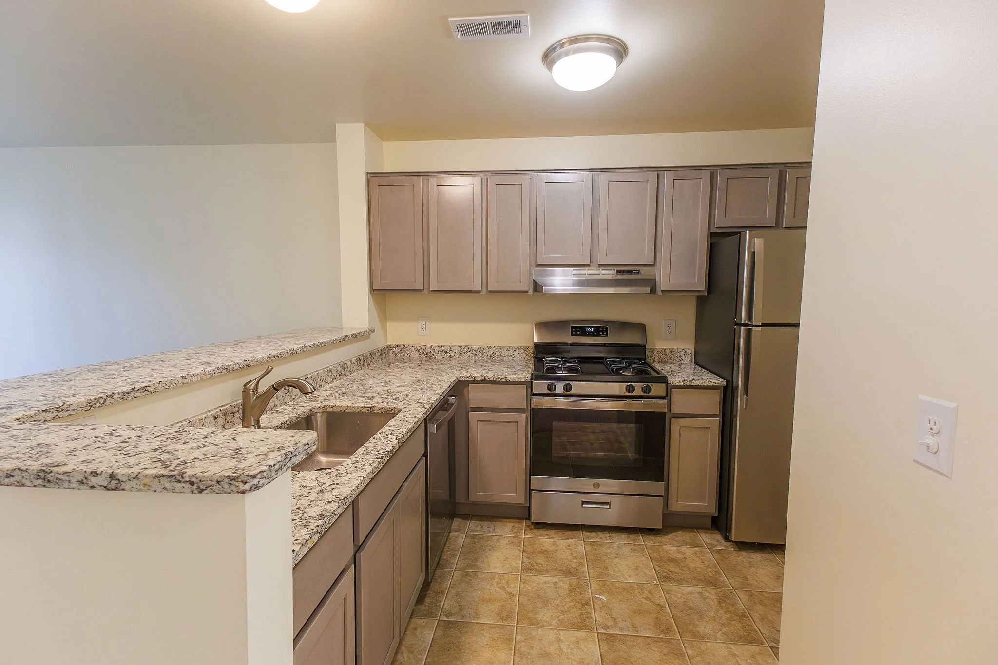 Kitchen with granite countertops, beige cabinets, stainless steel appliances including a refrigerator and stove, small sink, and tiled floor.