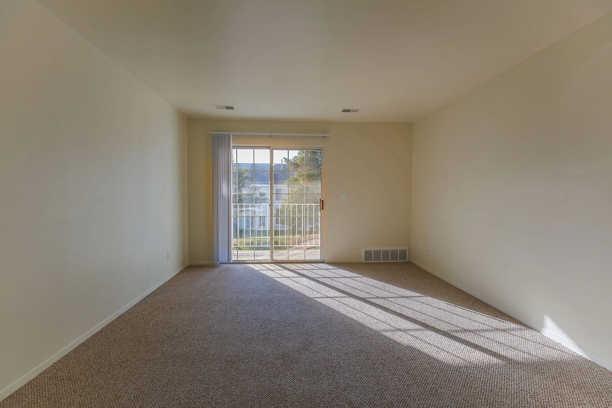 Empty living room with beige carpet, light yellow walls, sliding glass door with vertical blinds, sunlight casting shadows on the floor, and a view of a backyard with trees and houses.