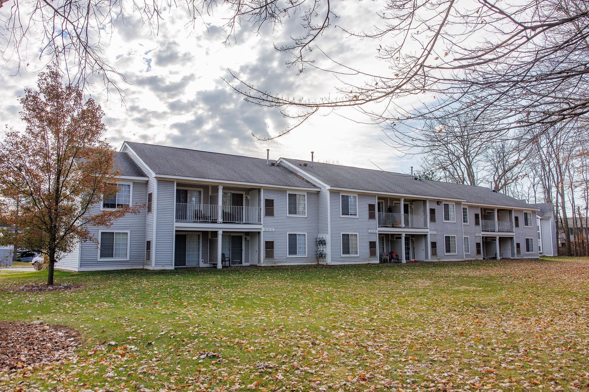 A two-story apartment building with white siding, multiple balconies, and a grassy yard with fallen leaves and a small tree in the foreground.