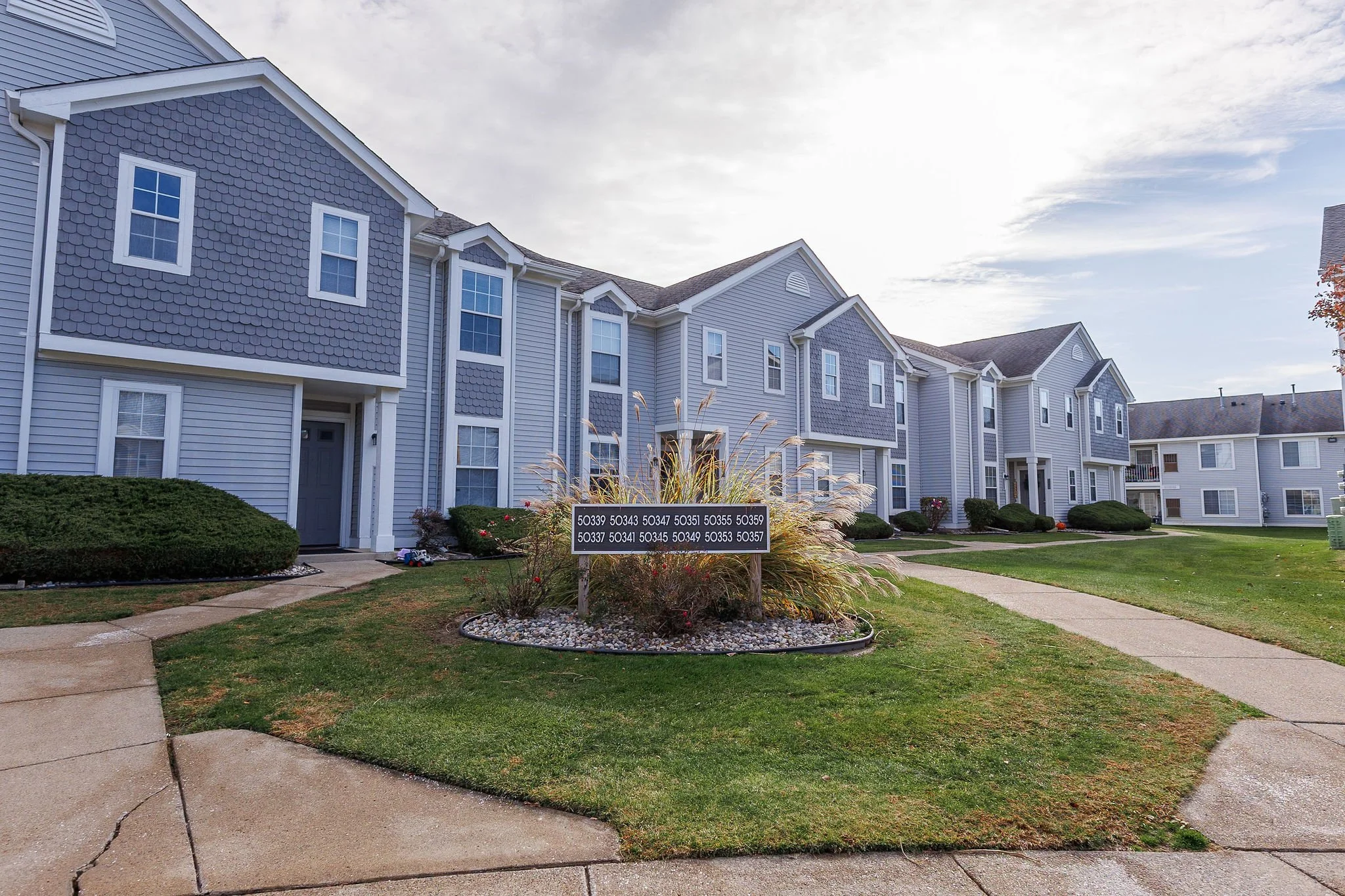 View of a residential apartment complex with multiple buildings, well-maintained lawn, a flower bed with a sign showing various number codes, and a sidewalk.