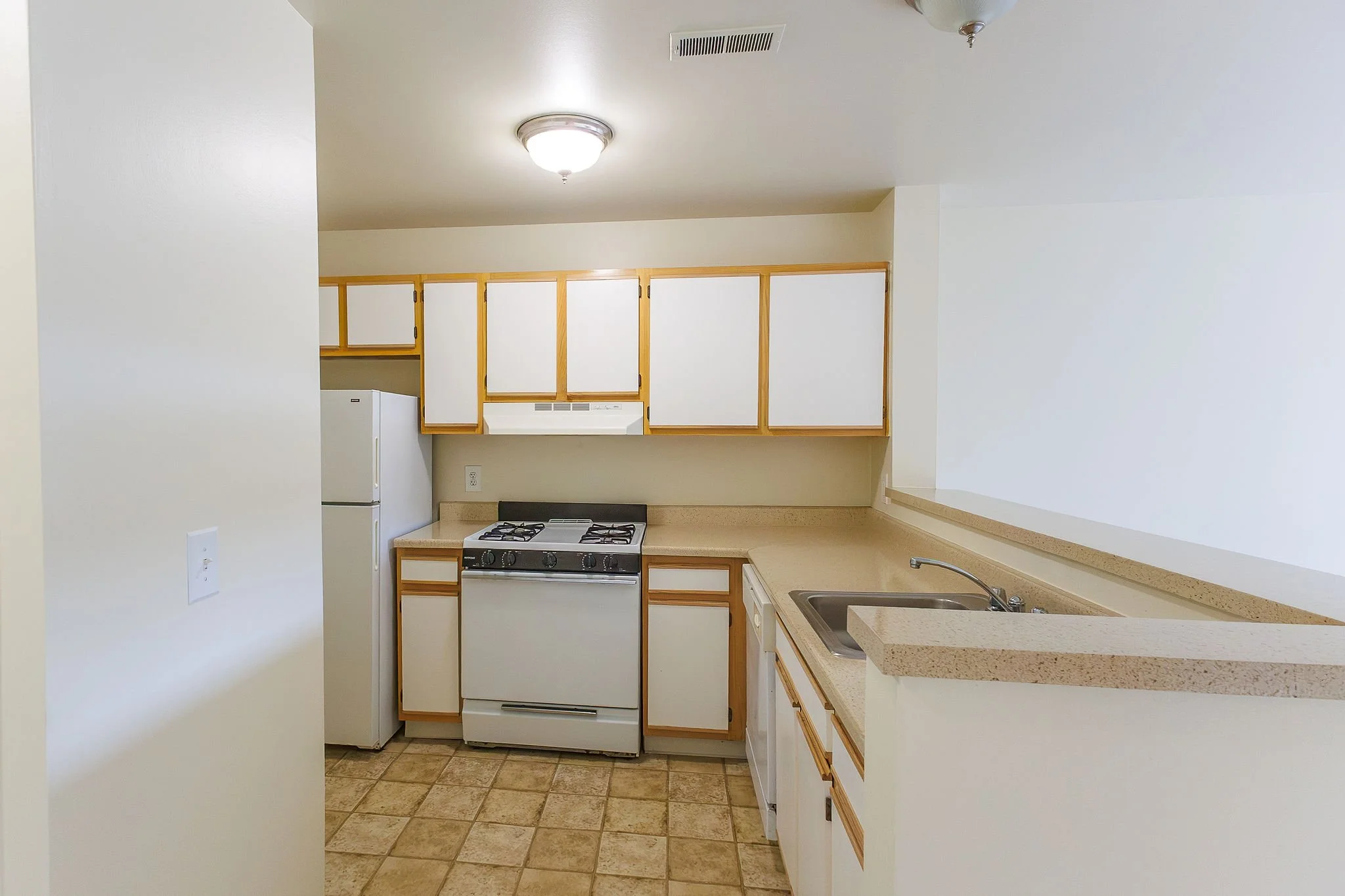 Empty kitchen with white cabinets, beige countertops, a white stove, refrigerator, and a stainless steel sink, featuring tile flooring and ceiling light fixtures.