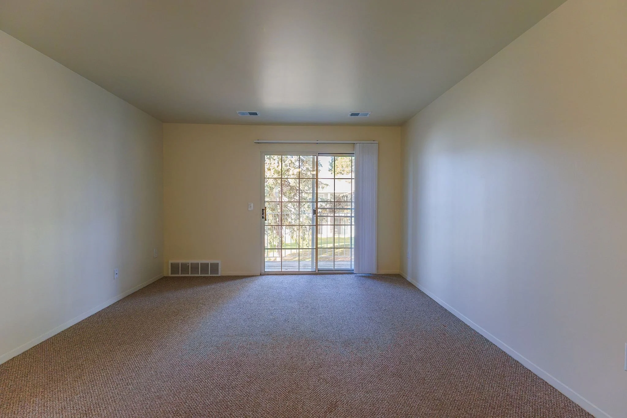 Empty living room with beige walls, carpeted floor, sliding glass door leading outside, vertical blinds, and a ceiling with vents.