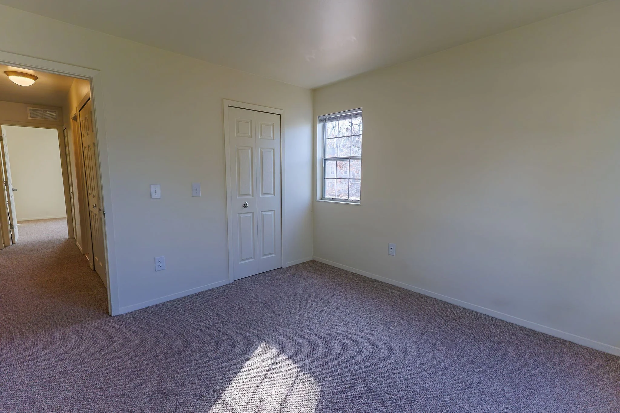 Empty bedroom with beige carpet, off-white walls, a window, a closet door, and a hallway entrance.