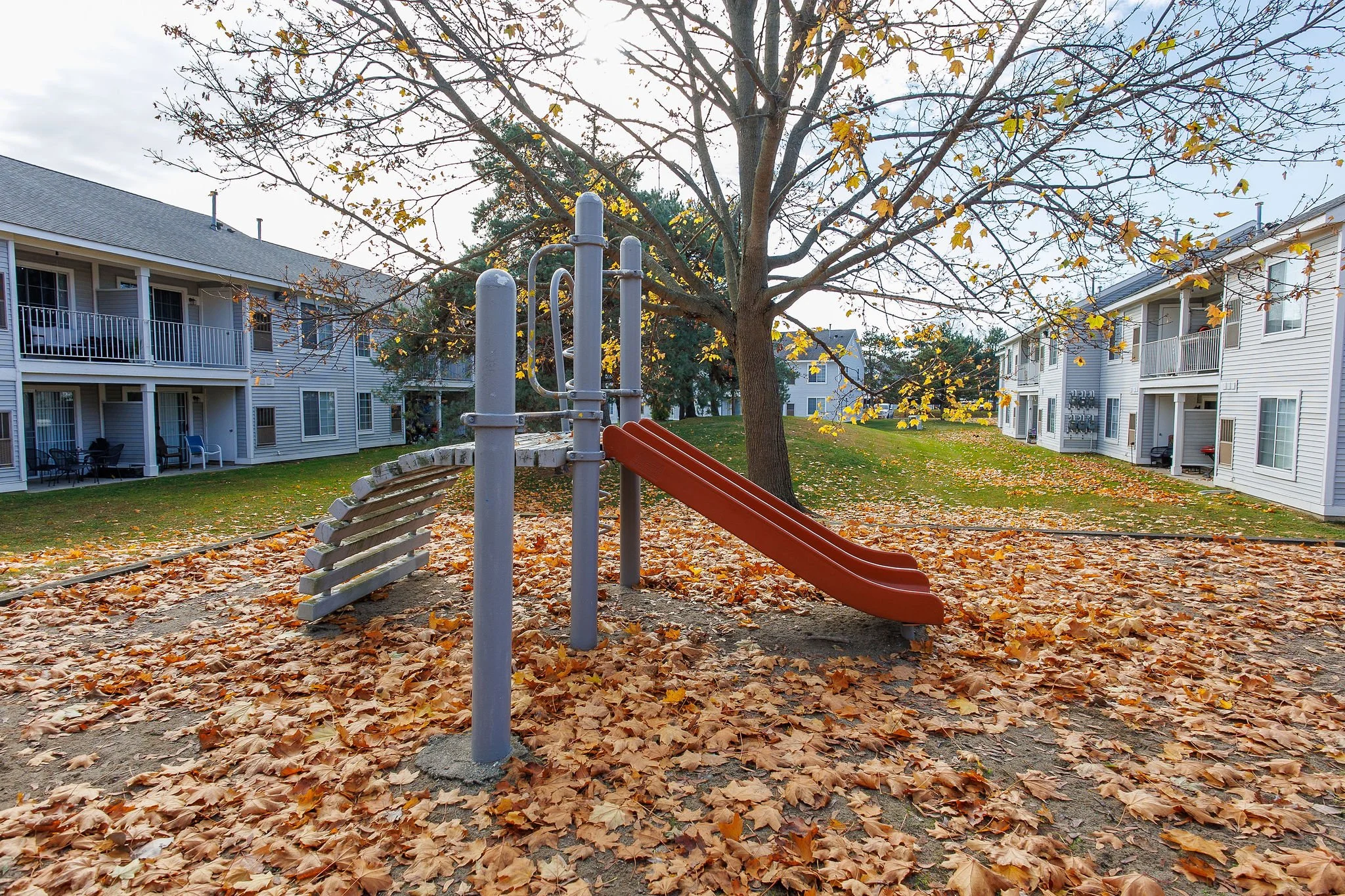 A playground slide and climbing ramp under a large tree with fall-colored leaves scattered on the ground in an apartment complex courtyard.