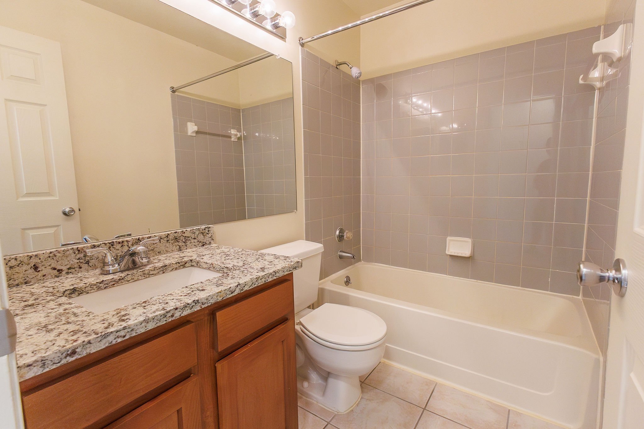 Bathroom with granite countertop sink, mirror, toilet, bathtub with gray tiled wall and soap holder.
