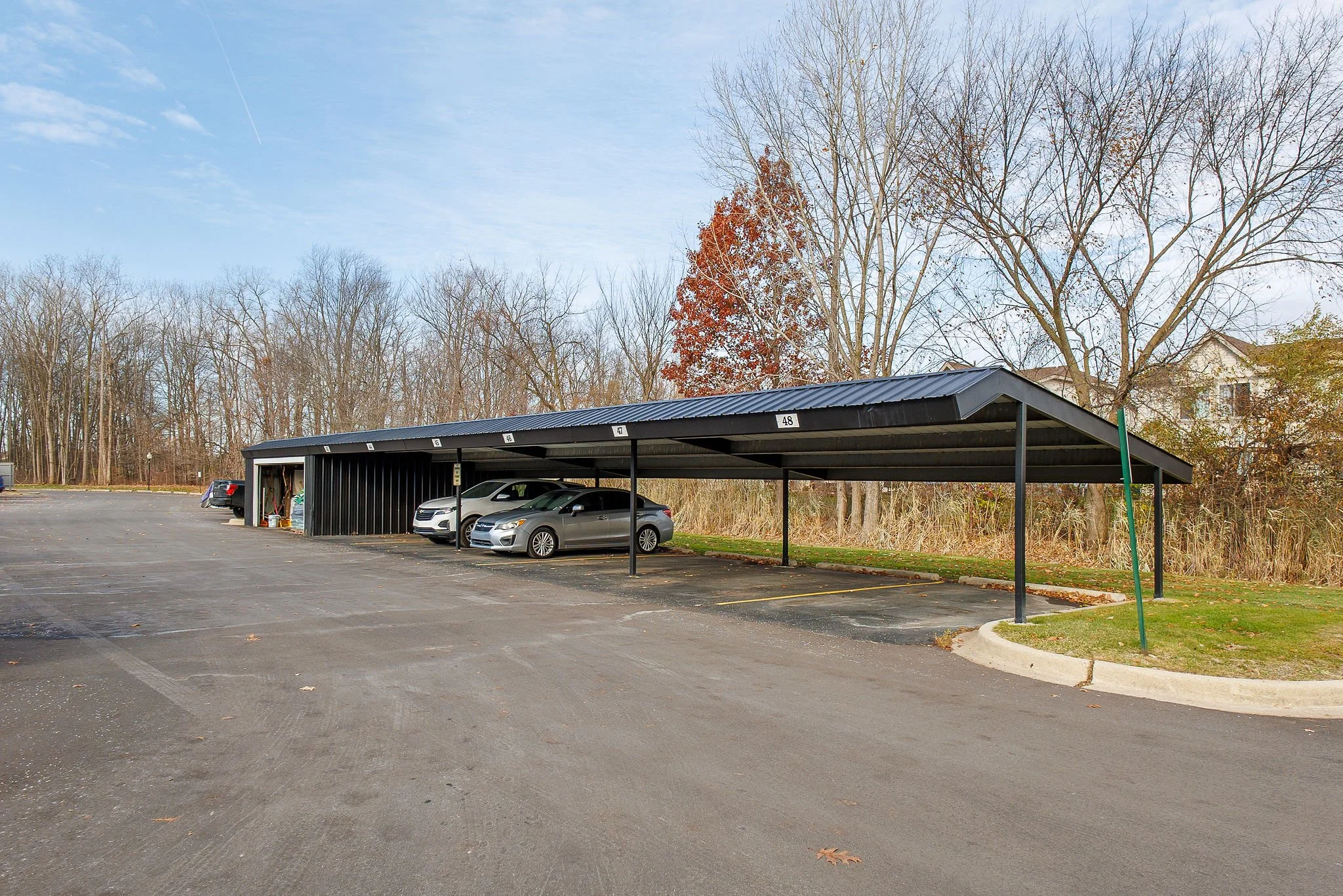 A parking lot with a covered carport structure and several parked cars. The background features leafless trees and a clear sky.