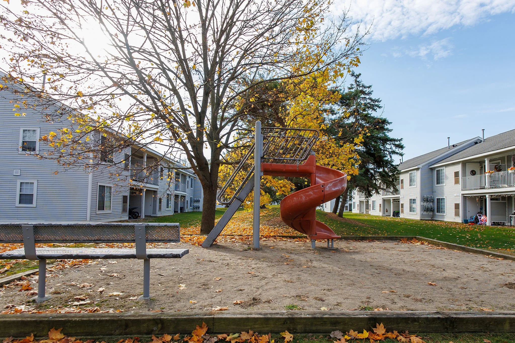 A playground slide in an apartment complex with residential buildings on either side, a tree with autumn leaves, and a bench nearby.