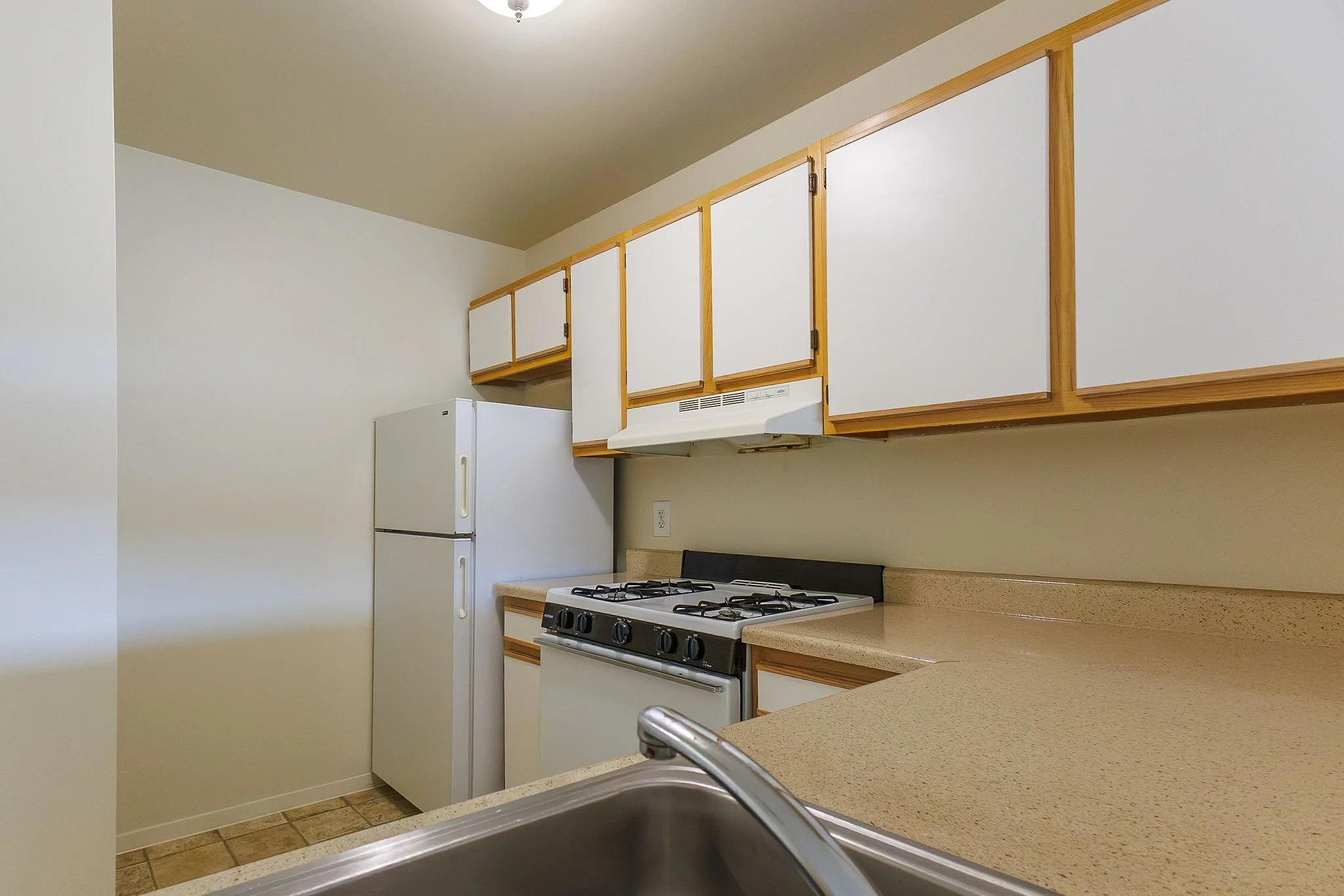 A kitchen with white upper cabinets, beige countertops, a white refrigerator, a black and white gas stove, and part of a stainless steel sink in the foreground.