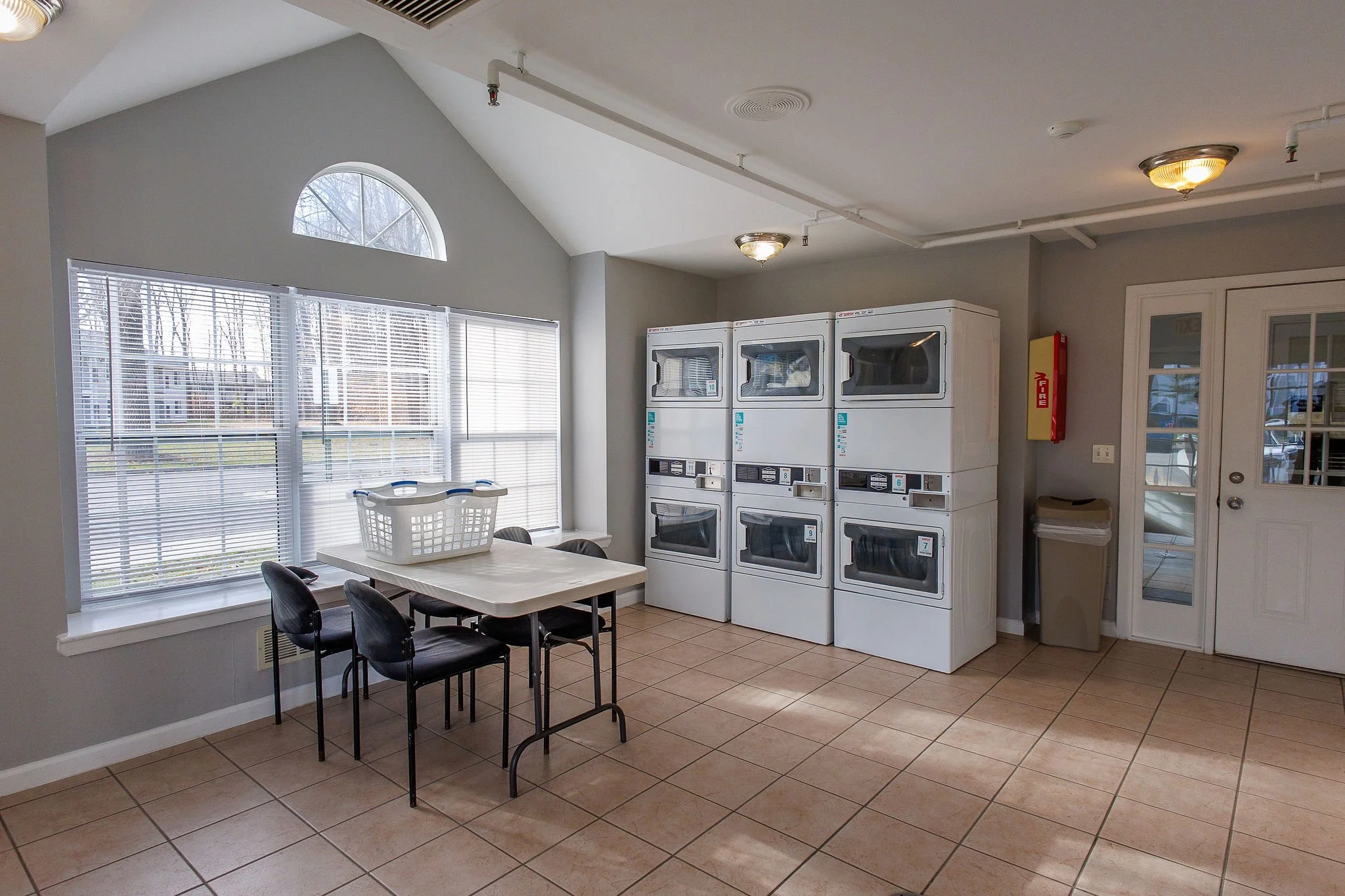 Laundry room with three washers, a table with chairs, laundry basket, large window, and a glass door.