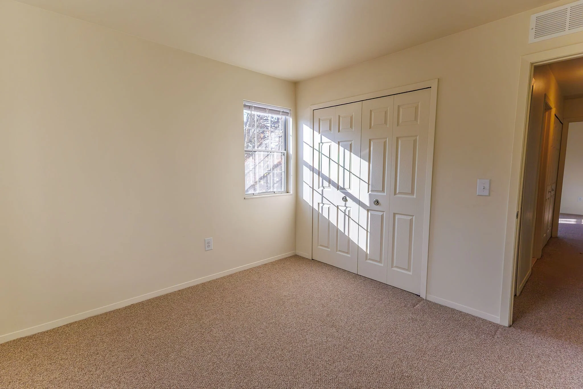 Empty room with beige walls, beige carpet, a window with sunlight and blinds, and a closed white closet with two doors.