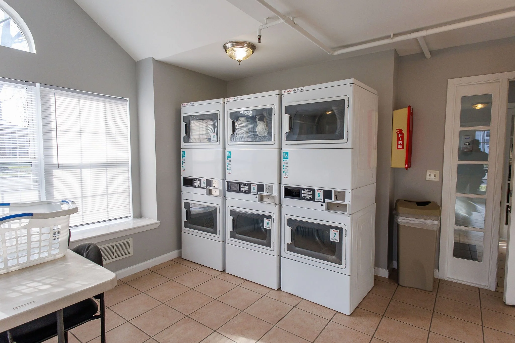 Laundry room with three stacked white dryers, a window with blinds, a table with a laundry basket, and a trash can near a fire alarm box.