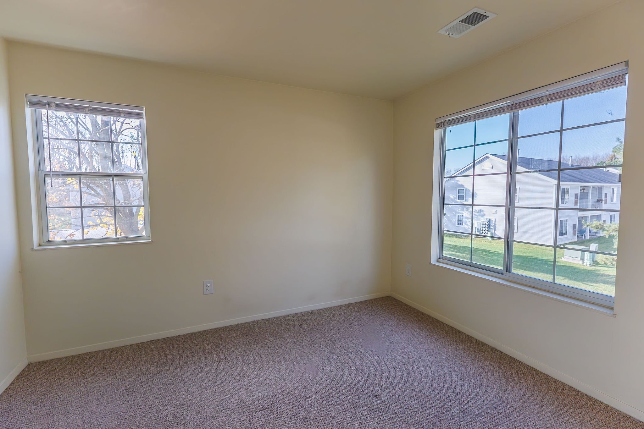 Empty room with cream walls, beige carpet, and large windows showing neighboring houses and trees.