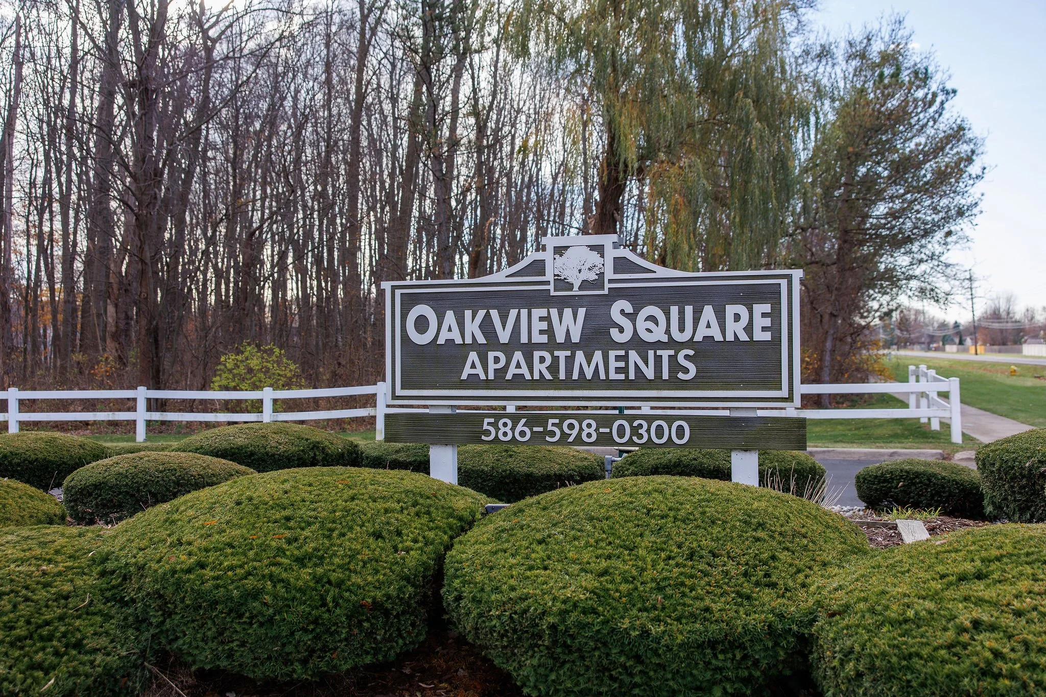 Sign for Oakview Square Apartments with phone number, surrounded by neatly trimmed bushes and a white fence, with leafless trees in the background.