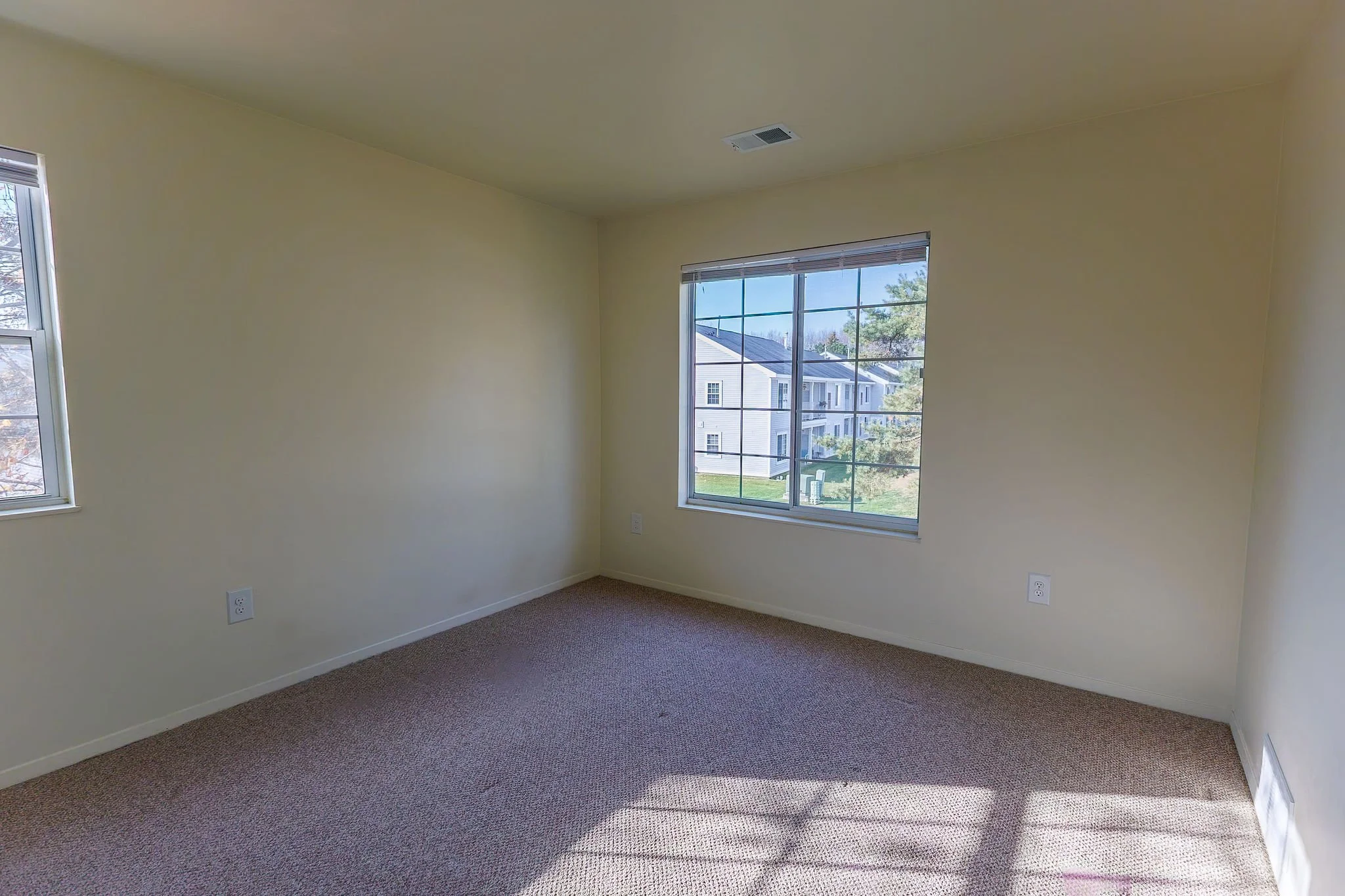 Empty room with beige walls, carpeted floor, two windows letting in natural light, and an air vent on the ceiling.