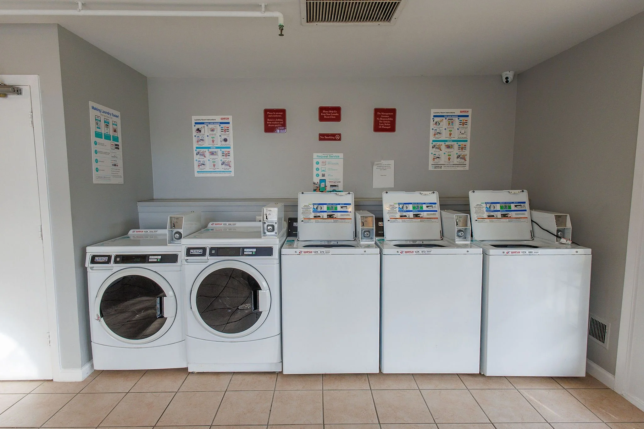 Coin-operated laundry machines including two front-loading washers and three top-loading dryers in a laundromat with informational and warning signs on the wall.