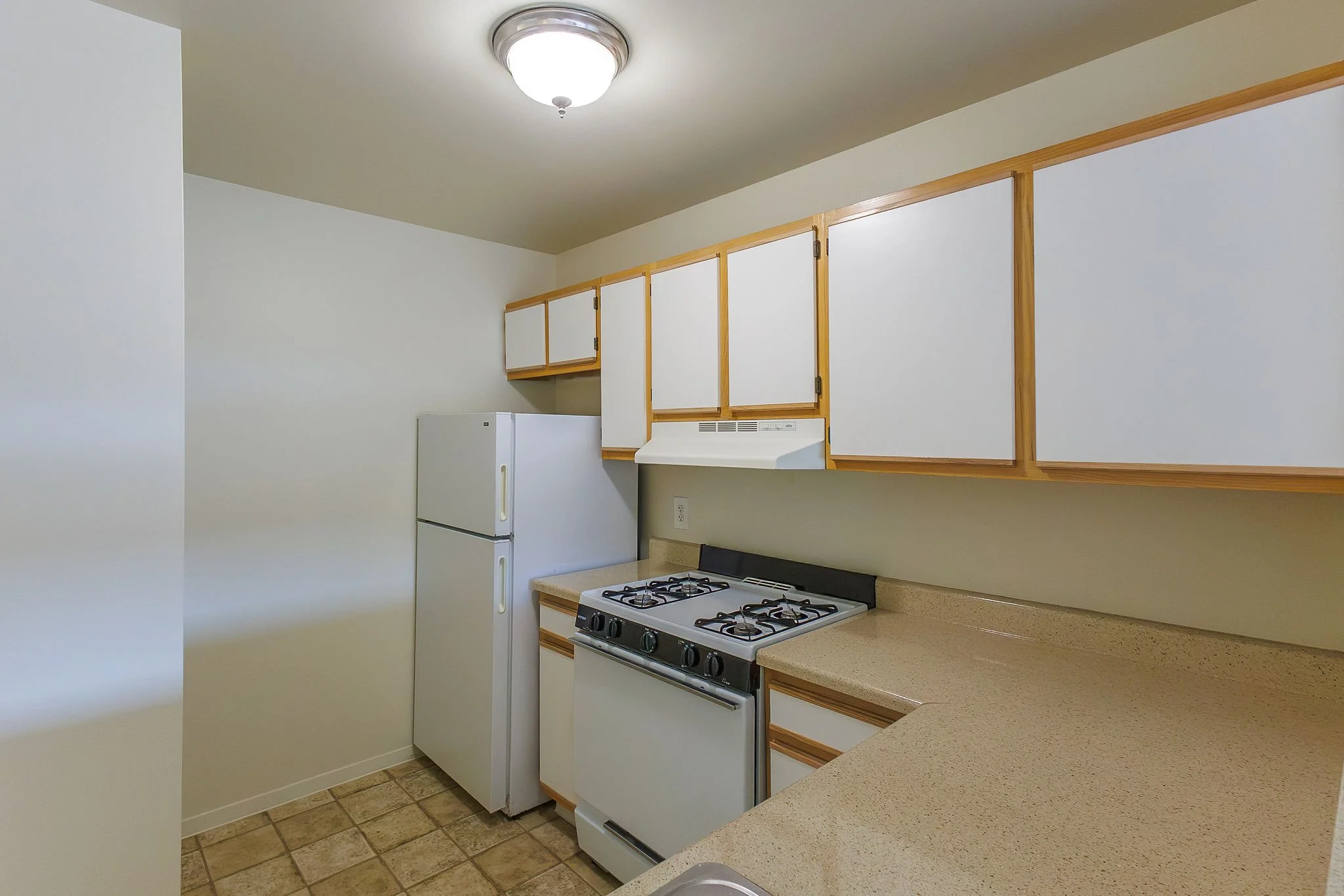 A small kitchen with white cabinets, a white refrigerator, a four-burner stove with an oven, beige countertops, a tiled floor, and a ceiling light fixture.