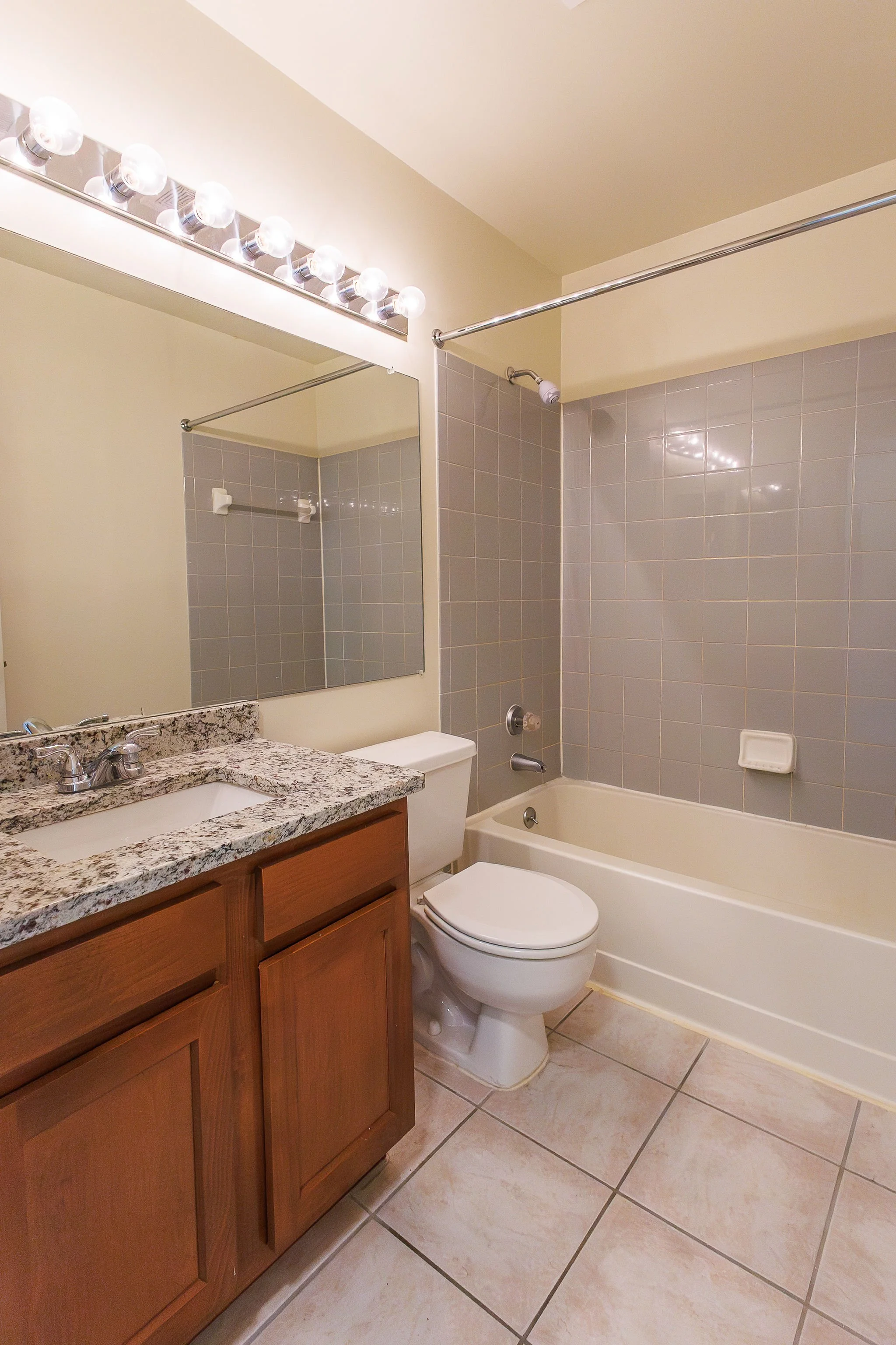 Bathroom with bathtub, toilet, wooden vanity with granite countertop, large mirror, tiled floor, and wall-mounted light fixture.