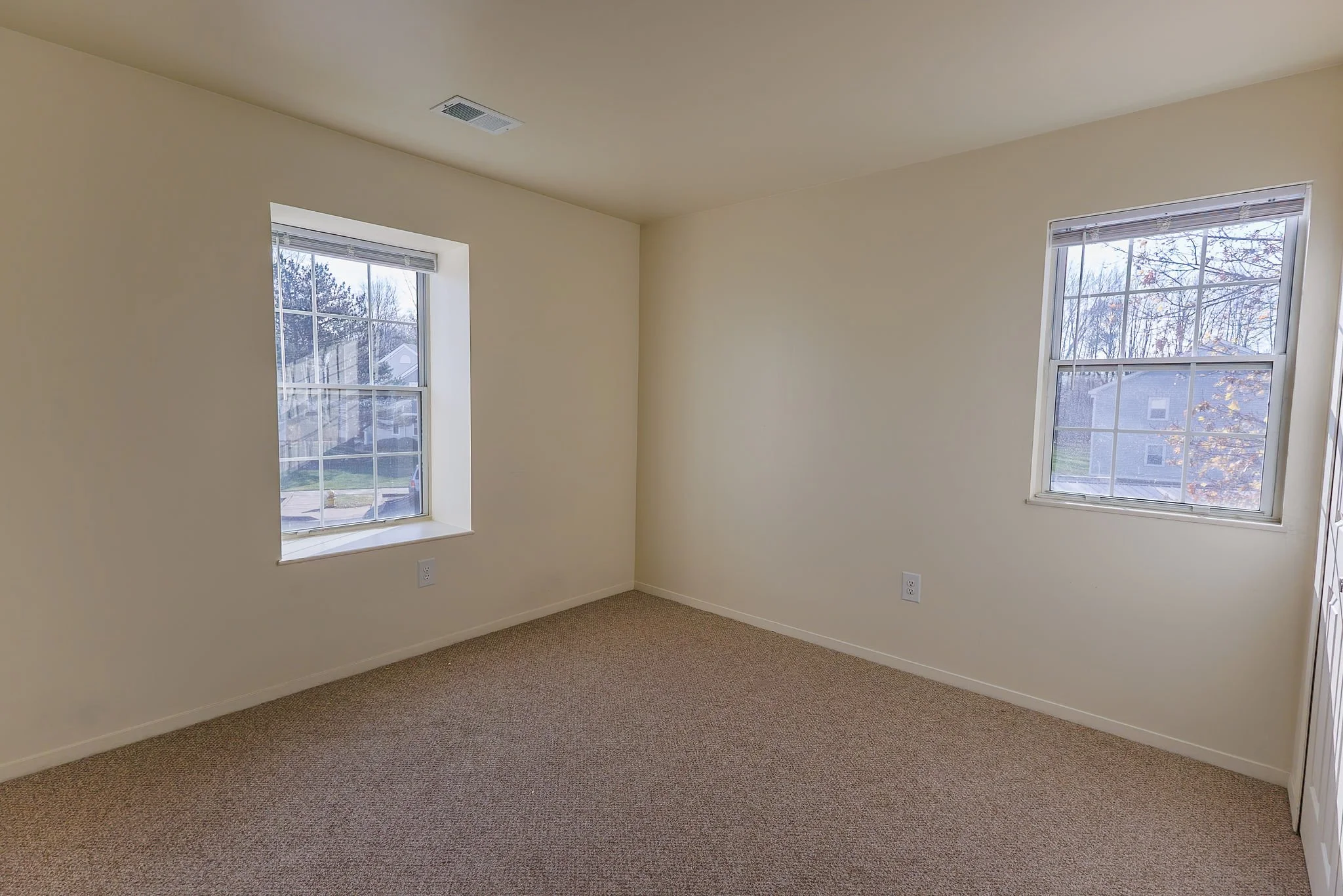 Empty room with beige walls, two windows showing a suburban neighborhood, and brown carpeting.