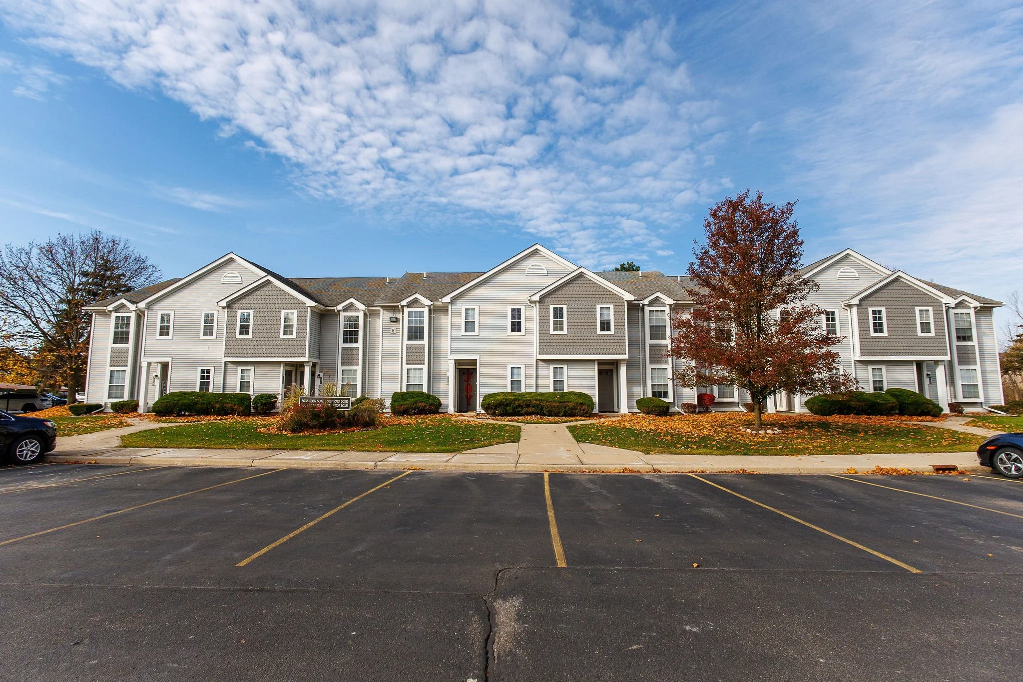 A multi-story residential apartment complex with white and gray exterior, surrounded by landscaped lawns, bushes, and trees, under a partly cloudy sky, with a parking lot in the foreground.