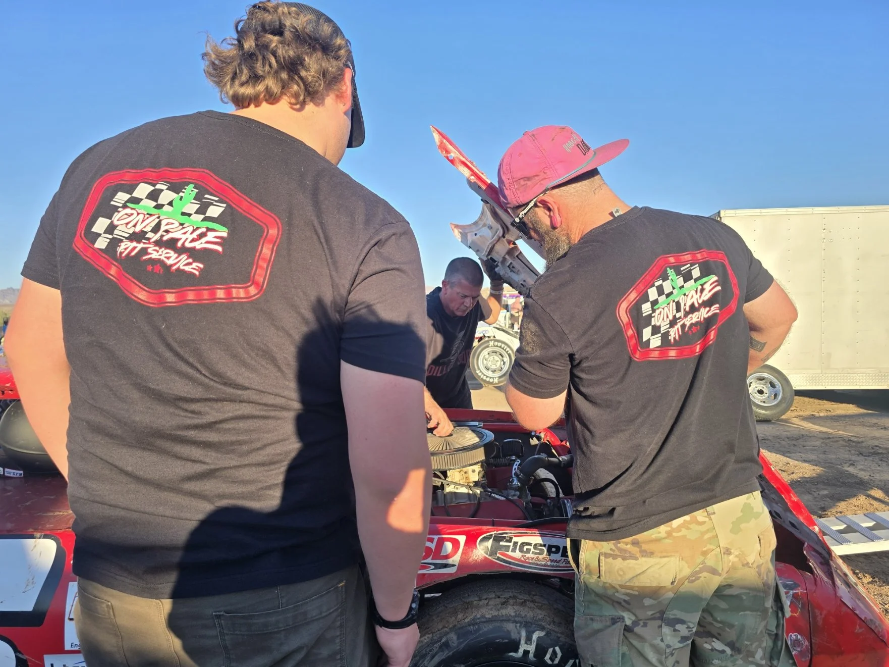 Three men are working on a red race car, with two of them closely inspecting the engine. They are outdoors on a sunny day, with a trailer visible in the background. The men are wearing black shirts with a logo and text that appears to say 'On Pace' a