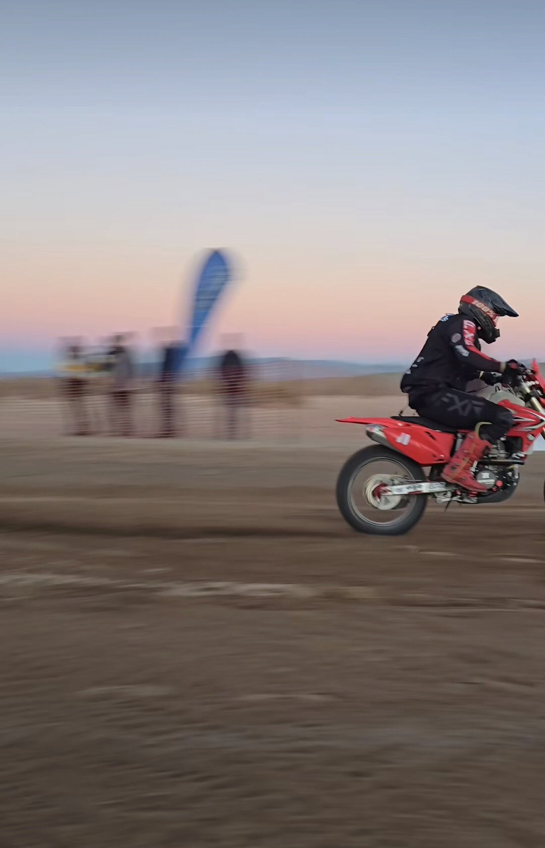 A motorcyclist in black riding gear and a full-face helmet riding a red dirt bike on dirt terrain during sunset, with blurred spectators and a blue flag in the background.