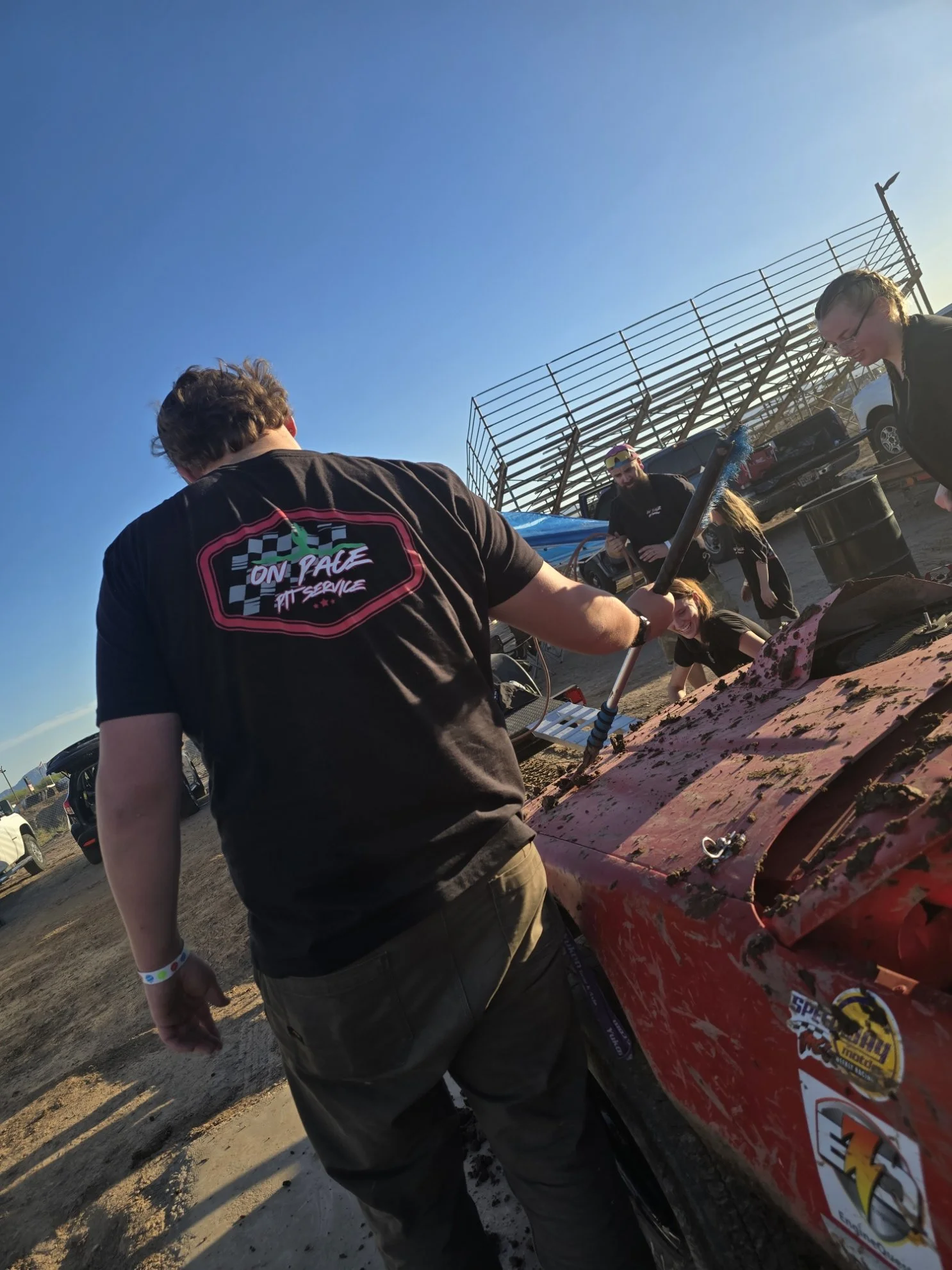 People working on a red race car at an outdoor event, with a metal bleacher and parked cars in the background on a clear day.