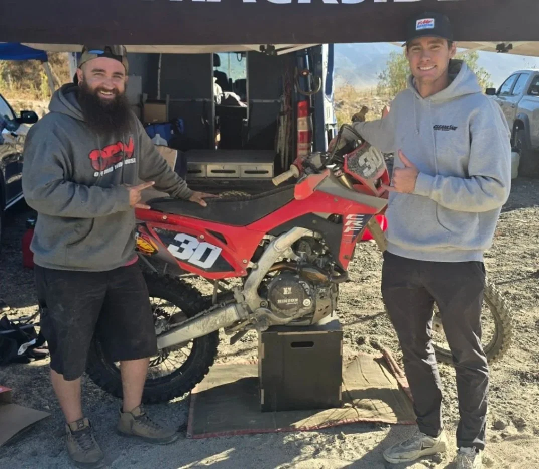 Two men standing next to a red and black dirt bike on a stand, outdoors with a truck and vehicles in the background, under a tent.