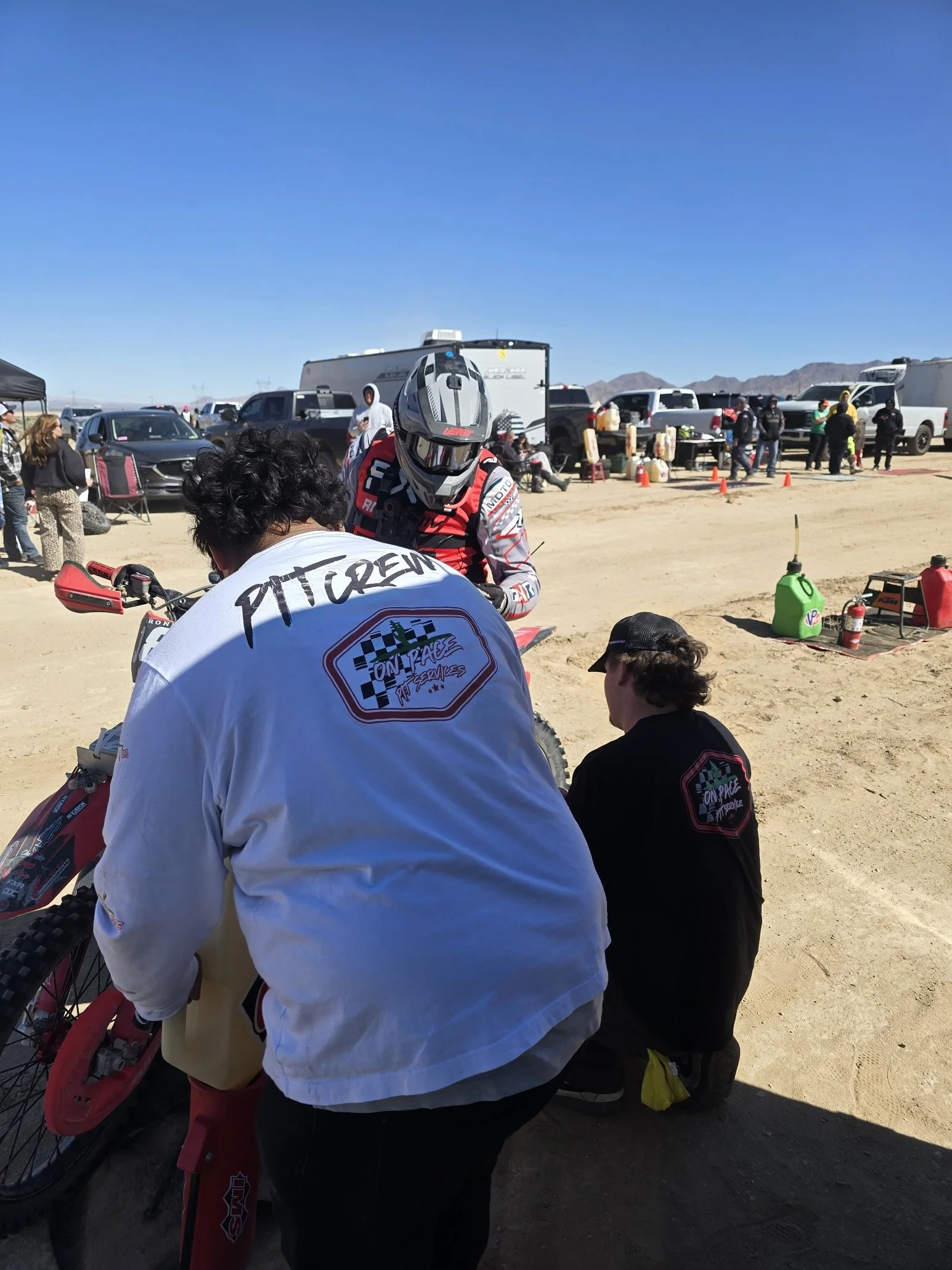 People preparing and getting ready for a dirt bike race in a desert area with parked vehicles and mountains in the background.