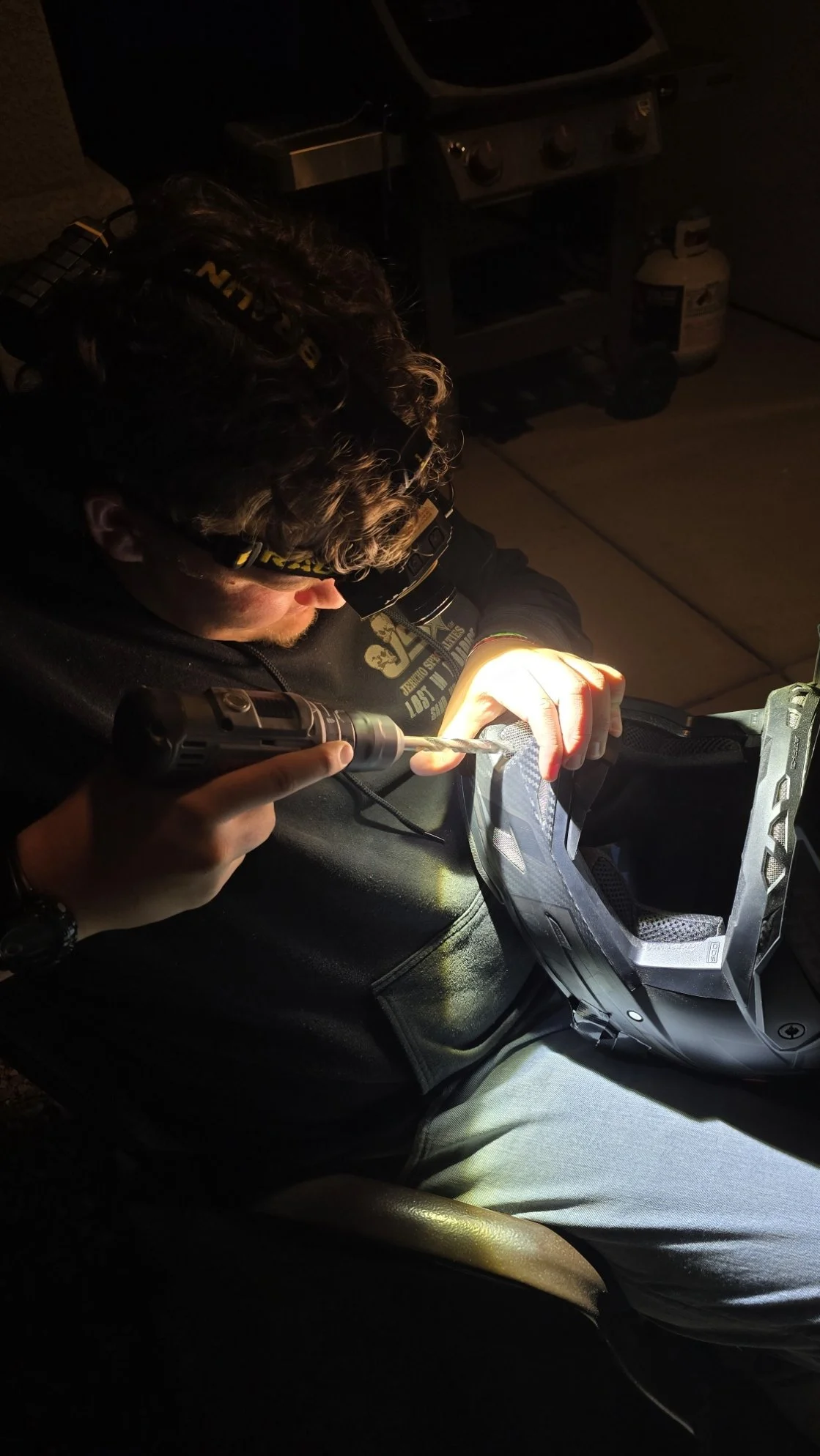 A person wearing a headlamp works on a motorcycle helmet with a tool inside a dimly lit garage.