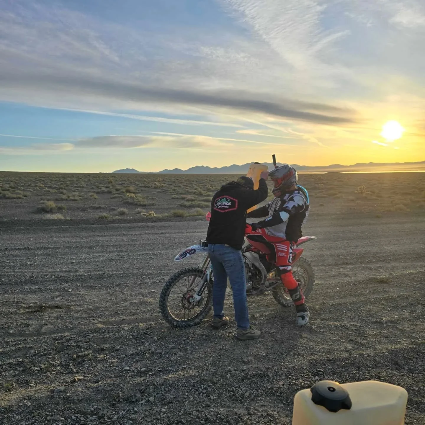 A motocross rider sits on a dirt bike while another person, dressed in a black hoodie, adjusts the rider’s helmet in a barren desert landscape during sunset, with mountains in the distance and a partly cloudy sky.