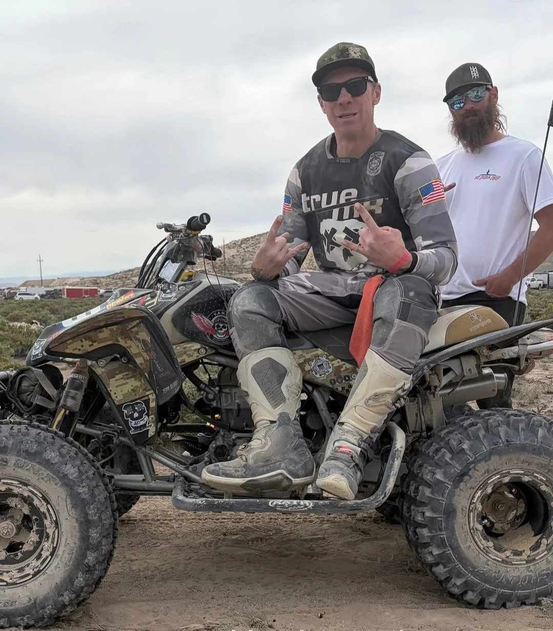 Two men on a dirt bike in a desert landscape, one is sitting and making rock hand gestures, wearing motocross gear, and the other is standing behind, wearing a baseball cap and sunglasses.