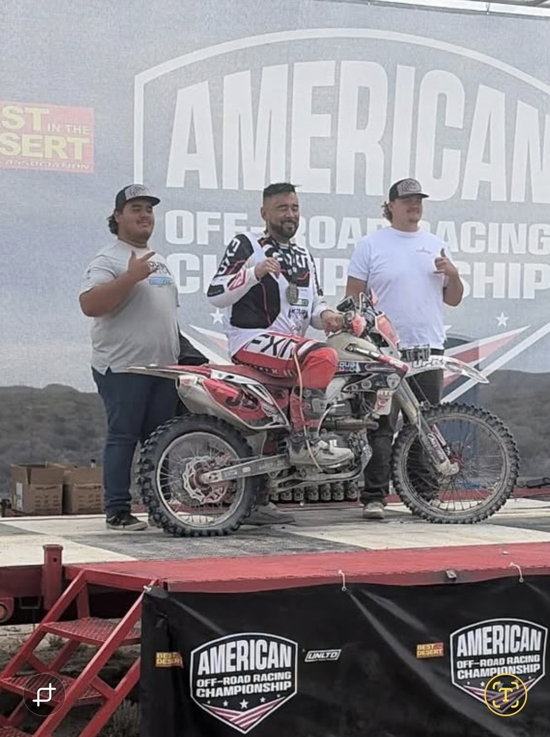 Three men standing on a podium with a dirt bike, celebrating at an off-road racing championship event. The background features a large sign that reads 'American Off-Road Racing Championship'.