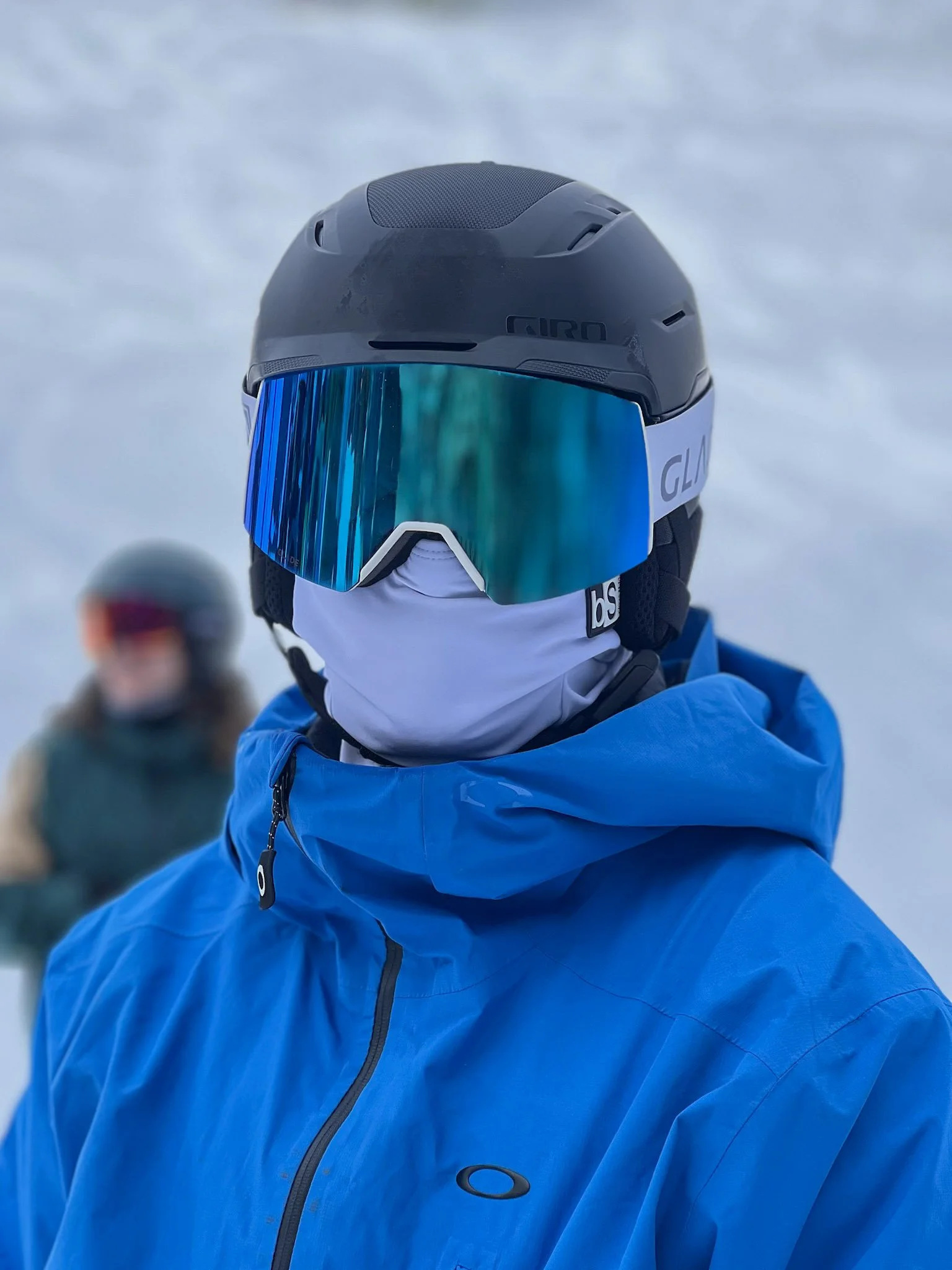 A person wearing a black ski helmet, blue polarized goggles, a white face mask, and a bright blue jacket standing in a snowy environment, with another person blurred in the background.