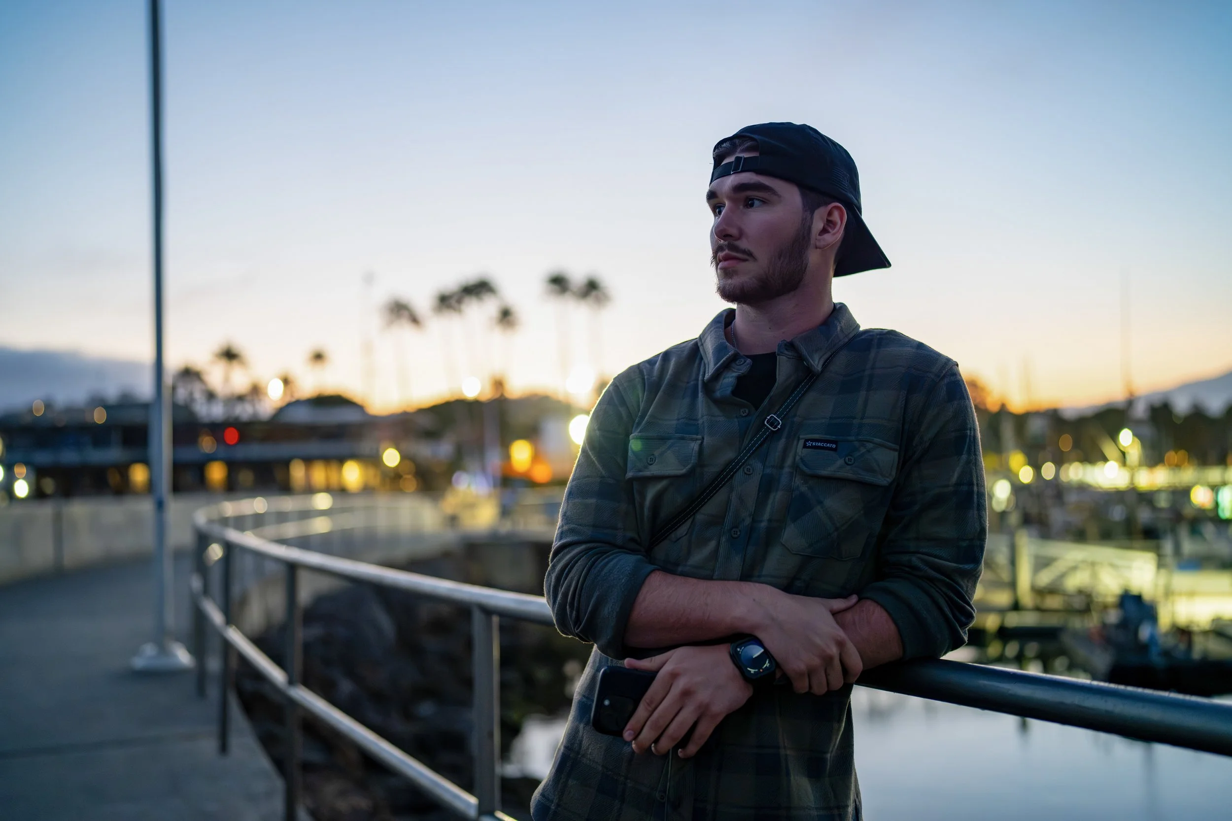 Young man with a beard, wearing a backward baseball cap, plaid shirt, and smartwatch, standing by a waterfront railing during sunset, with palm trees and city lights in the background.