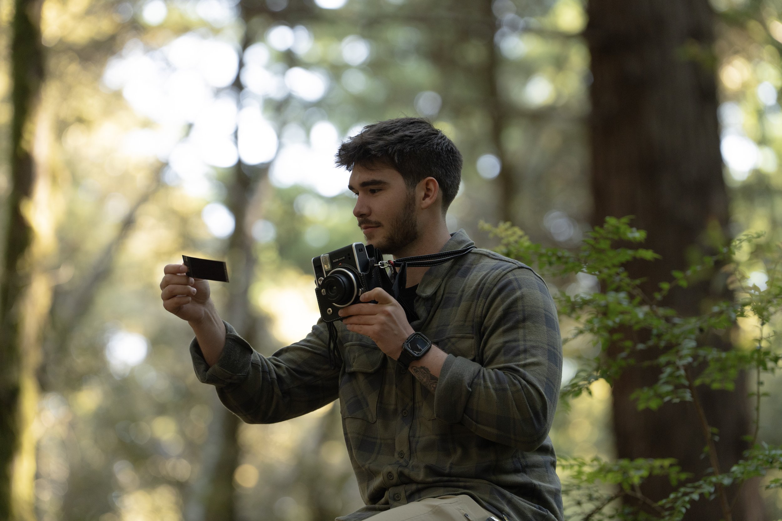 A young man with a camera looking at a photograph on a card, sitting outdoors in a forest with sunlight filtering through trees.