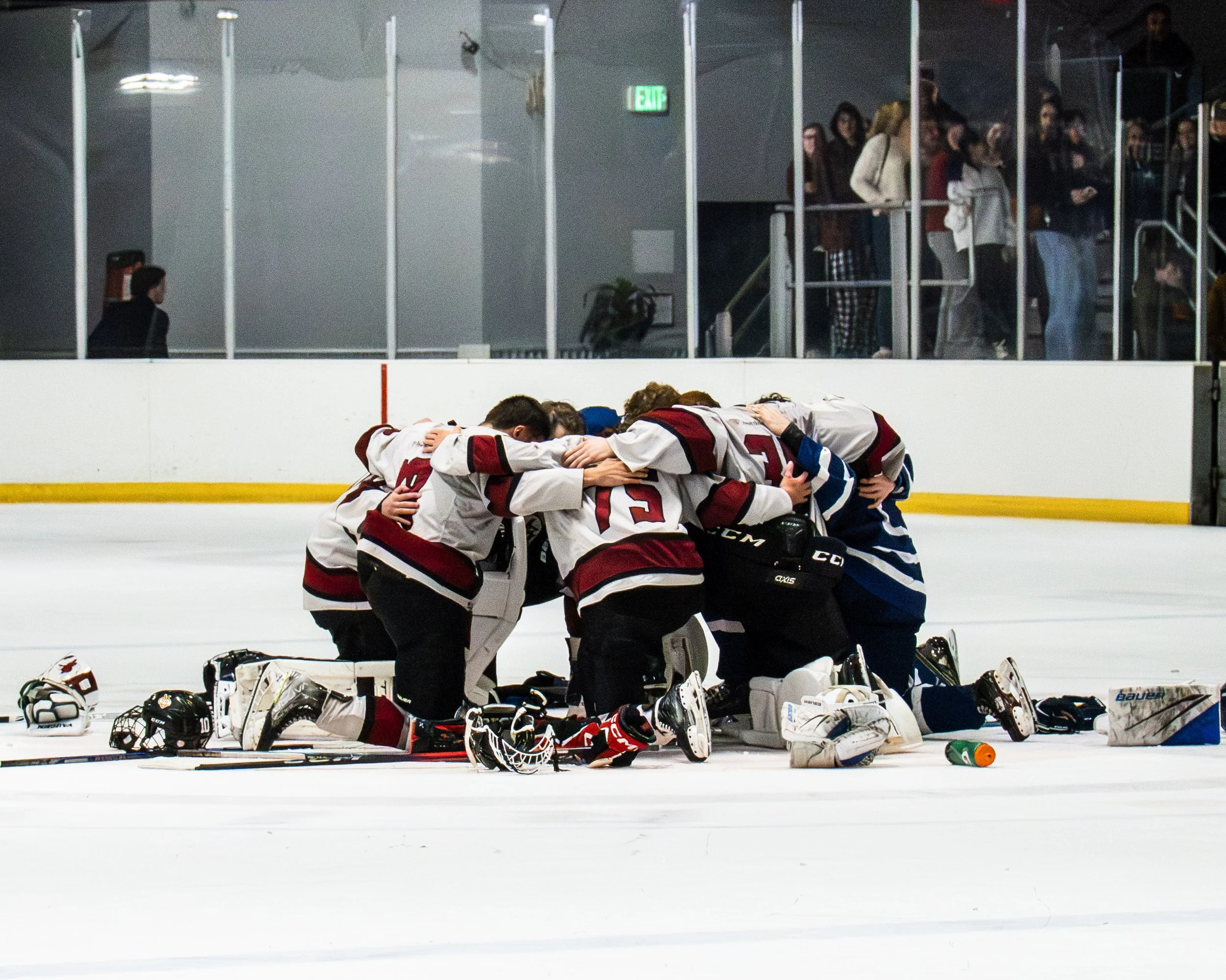 Ice hockey team huddled together in a team prayer or celebration on the ice rink, with helmets and gear scattered around.