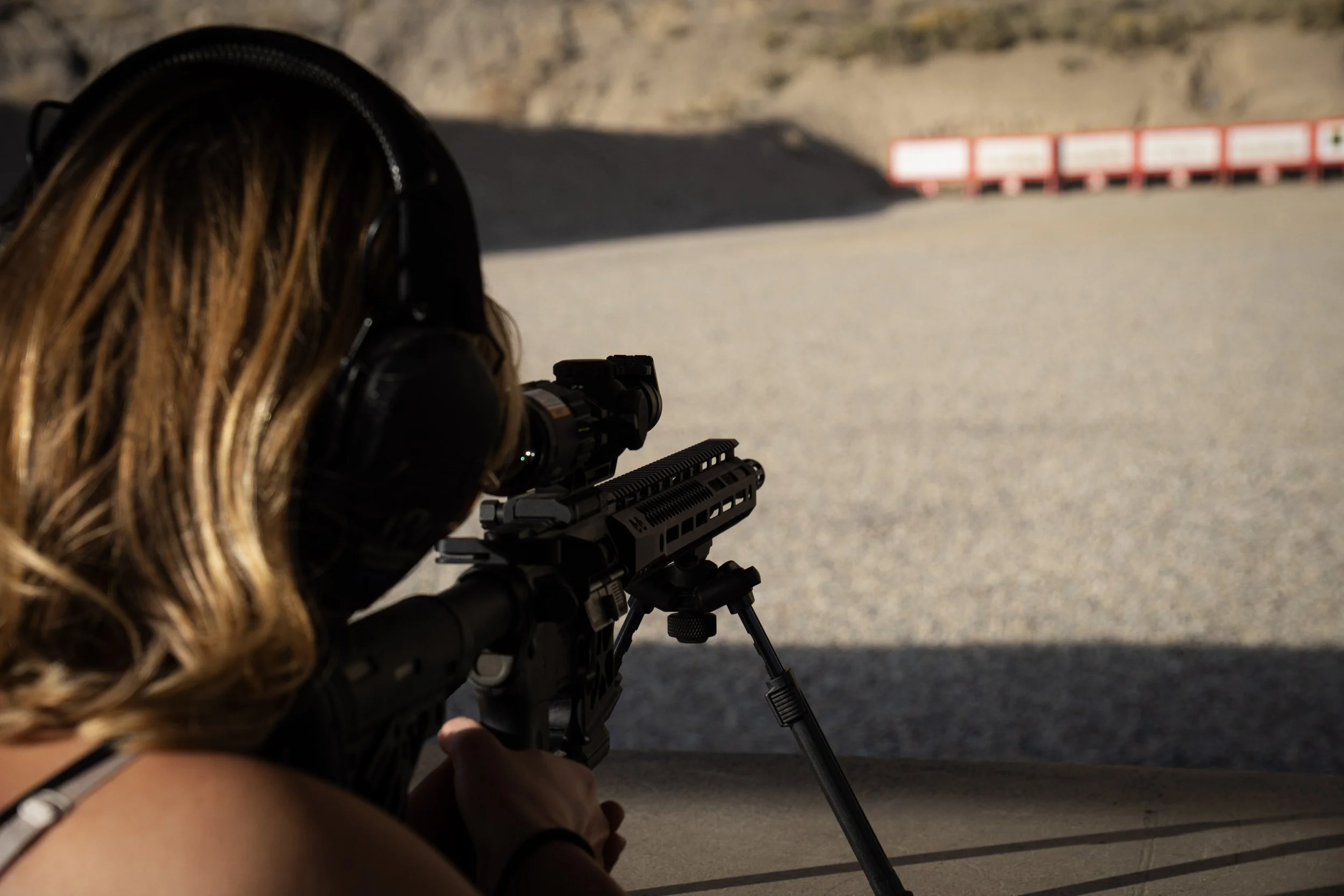 A woman with long, wavy blonde hair, wearing safety headphones, aiming a scoped rifle on a bipod at an outdoor shooting range.