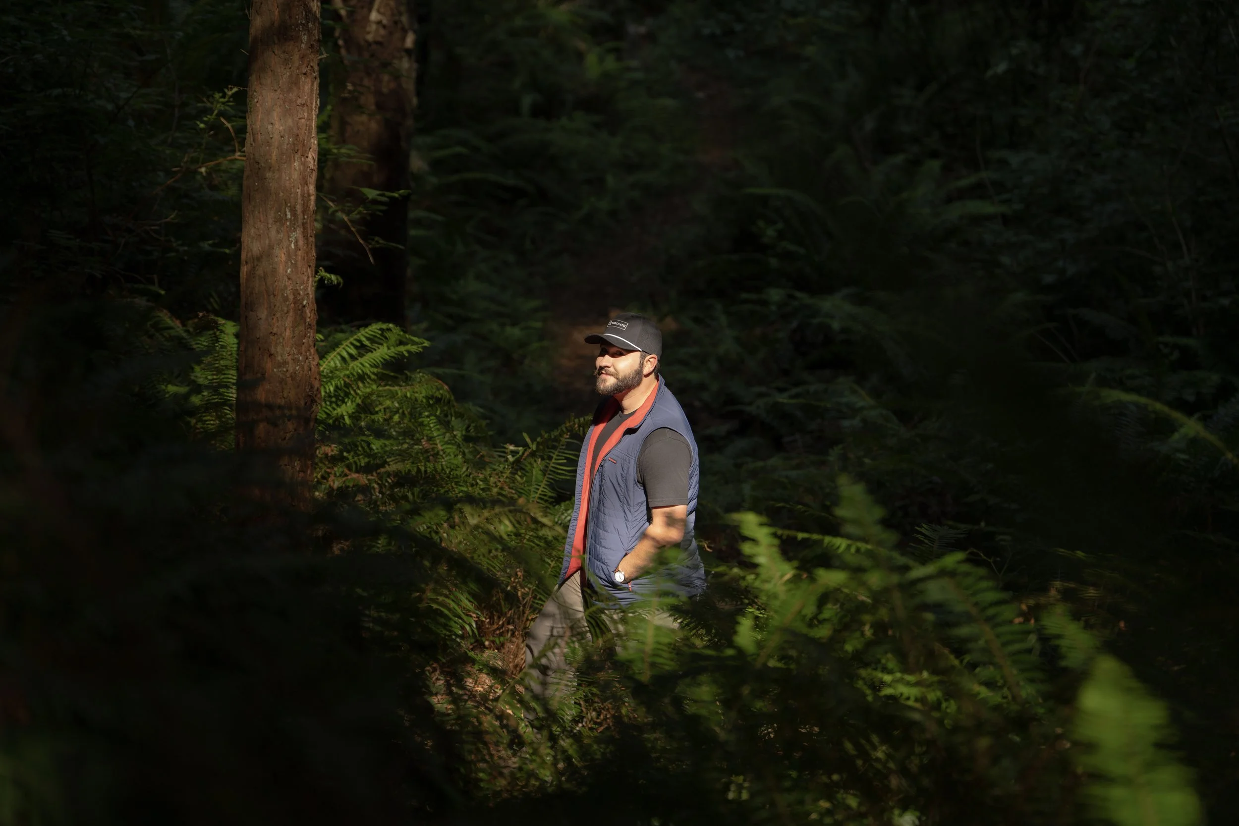 A man with a beard wearing a black cap, black t-shirt, and blue vest standing in a lush, dense forest with sunlight filtering through the trees.