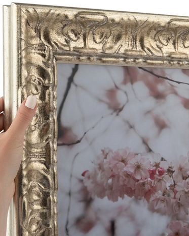 Close-up of a hand with a white nail holding the corner of an ornate, vintage-style gold picture frame with floral artwork inside.