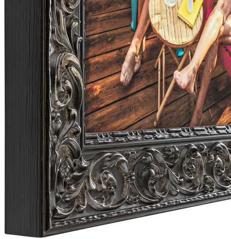 Close-up of an ornate black-framed mirror reflecting a wooden deck with a person sitting on a chair and a small round table with a yellow notepad and a yellow watch.