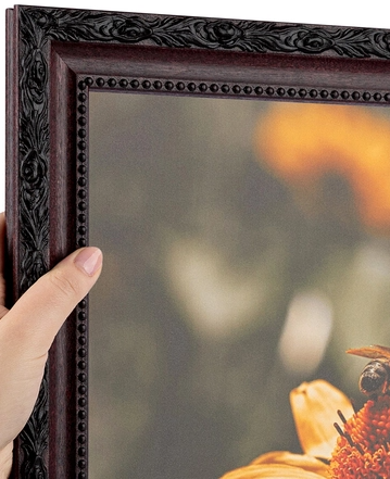 A person is holding a decorative framed mirror with a black ornate border, reflecting a blurred outdoor scene.