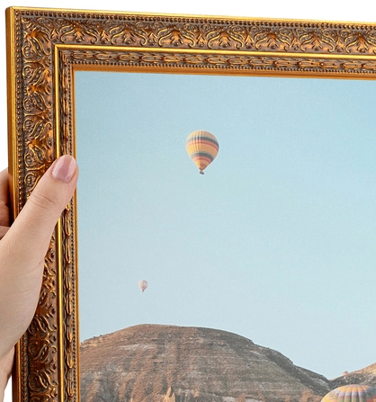 A person's hand holding a decorative gold picture frame, with hot air balloons floating in the sky visible through the frame.