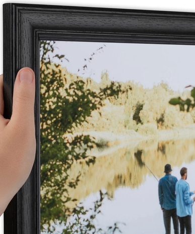 A person holding a picture frame showing a scene of two men fishing by a river with trees in the background.