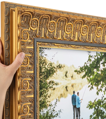 Person holding the corner of a decorative gold-colored picture frame with an ornate pattern, viewing a landscape painting of two people fishing by a river under trees.