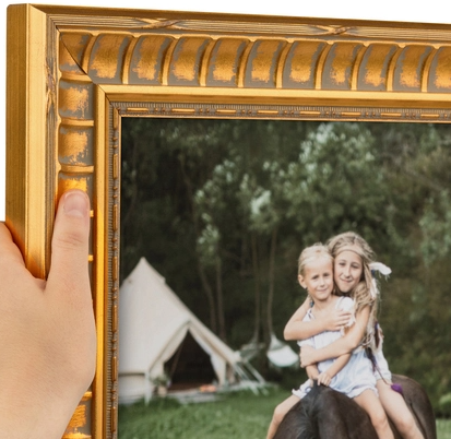 A hand holding an ornate gold picture frame around a photo of two young girls, one riding a horse and both smiling with arms around each other, outdoors with green trees and tents in the background.