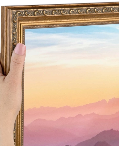 A hand holding a framed picture of a colorful sunset or sunrise landscape with mountains and a sky in pastel shades.
